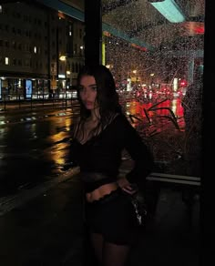 a woman standing in front of a window with rain falling down on her and buildings lit up at night