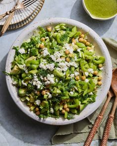 a white bowl filled with green vegetables and feta cheese next to two wooden spoons