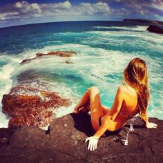 a beautiful woman sitting on top of a cliff next to the ocean with her legs crossed