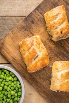 three pastries sitting on top of a wooden cutting board next to peas and a bowl