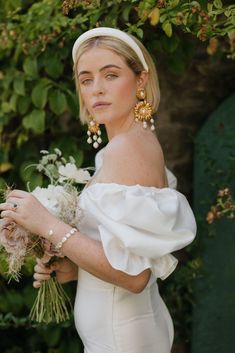 a woman in a white dress is holding flowers and wearing gold earrings on her head