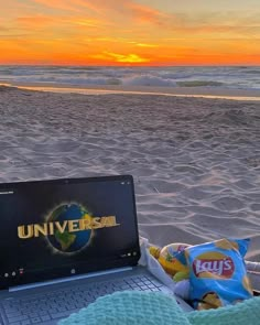 an open laptop computer sitting on top of a sandy beach next to the ocean at sunset