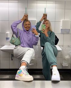 two women taking selfies in the bathroom with their cell phones and holding up cups