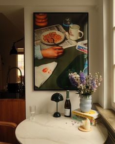 a painting hangs on the wall next to a table with flowers in it and books