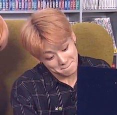 a man with blonde hair sitting in front of a book shelf and looking at his laptop