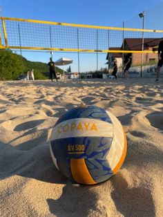 a volley ball sitting in the sand at a beach volleyball court with people playing on the sidelines