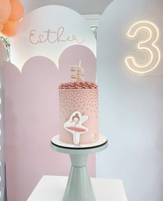 a pink and white birthday cake on a table in front of a wall with balloons