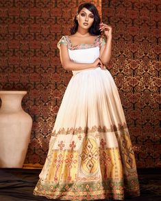 a woman standing in front of a wall wearing a white and gold dress with an intricate pattern