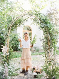 a woman standing in front of an archway with greenery and flowers on it,