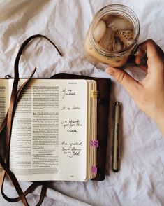 an open book with writing on it next to a hand holding a pen and a glass jar filled with rocks