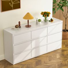 a white dresser sitting on top of a hard wood floor next to a lamp and potted plant