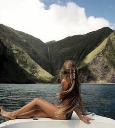 a beautiful woman sitting on top of a boat in the middle of water with mountains behind her