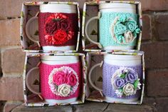 four coffee mugs decorated with crocheted flowers and ribbons are sitting on a piece of wood