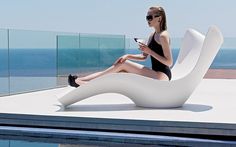 a woman sitting on top of a white chair next to a pool and looking at her cell phone