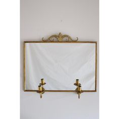 two brass candle holders in front of a white wall with a gold framed mirror on it