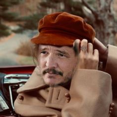 a man wearing a brown hat and coat sitting in a car with his hand on the steering wheel