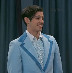 a young man in a blue suit and white shirt smiles at the camera while standing next to a curtain