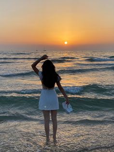 a woman standing in the ocean at sunset with her back to the camera and arms behind her head