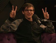 a man sitting in front of a microphone making the peace sign with his hands and wearing glasses