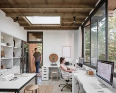 two people working in an office area with large windows and wooden ceilinging, along with white desks