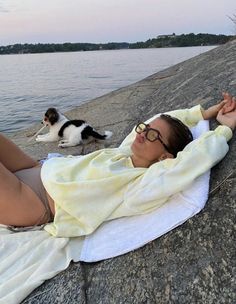 a woman laying on top of a rock next to a lake with a dog standing behind her