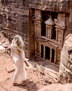 a woman in a white dress is walking through the desert near a rock formation and some buildings