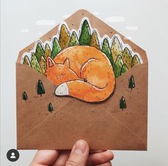 a hand holding up an envelope with a drawing of a sleeping fox in the woods