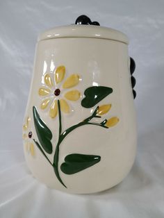 a white vase with yellow flowers and green leaves painted on the side, sitting on a white surface