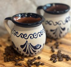 two coffee mugs sitting on top of a wooden table with coffee beans scattered around them