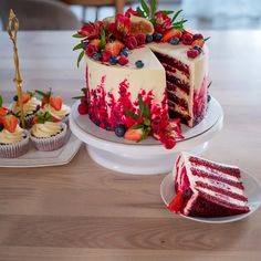 a red velvet cake with white frosting and fresh berries on top, surrounded by cupcakes