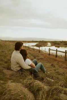 a man and woman sitting next to each other on top of a grass covered hill