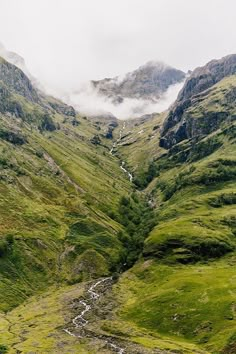 the valley is surrounded by green mountains and clouds in the distance, with a stream running between them