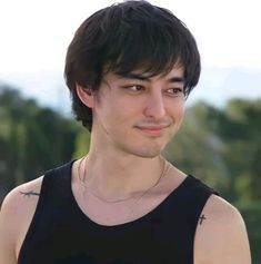 a young man wearing a black tank top standing in front of trees