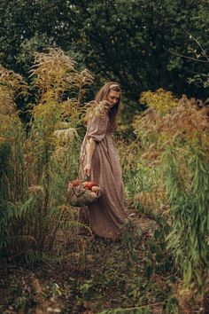 a woman in a long dress holding a basket full of apples walking through tall grass