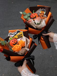 two hands holding orange and brown paper wrapped bouquets with halloween decorations on them,