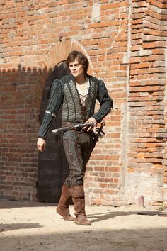 a young man dressed in medieval clothing standing next to a brick wall with his hands on his hips