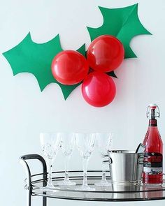 a christmas decoration with red balloons and holly leaves on the wall, next to wine glasses