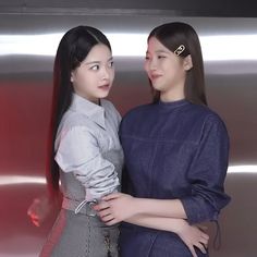 two young women standing next to each other in front of a metal wall and looking at the camera