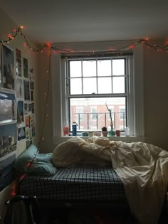 an unmade bed in front of a window with christmas lights strung across the room