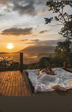 a woman laying on top of a wooden deck under a cloudy sky at sunset with the sun setting