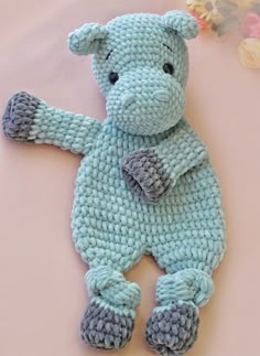 a crocheted teddy bear laying on top of a pink surface with flowers in the background