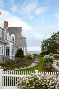 a white picket fence in front of a house with flowers on the lawn and water behind it