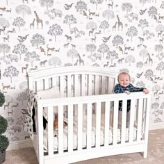 a baby sitting in a white crib next to a wall with animals on it