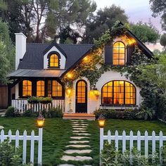 a white house with lots of windows and lights on it's front door, surrounded by greenery