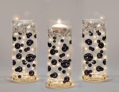 three glass vases with lights in them on a white surface, one is filled with black balls