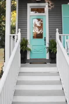 a blue door is on the side of a gray house with white stairs and potted plants