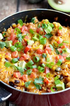 a skillet filled with rice and vegetables on top of a wooden table next to an avocado