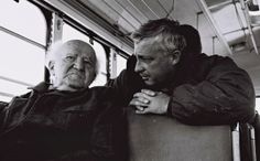 two elderly people sitting on a bus looking at something in the distance with their arms around each other