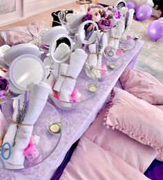 a purple table topped with lots of white plates and bowls filled with food next to pink pillows