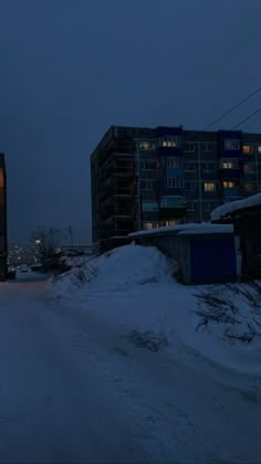 a truck driving down a snow covered road in front of tall buildings at night time